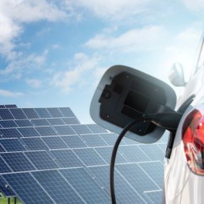 The side of an electric car, plugged in to a charging outlet with solar panels in the background.